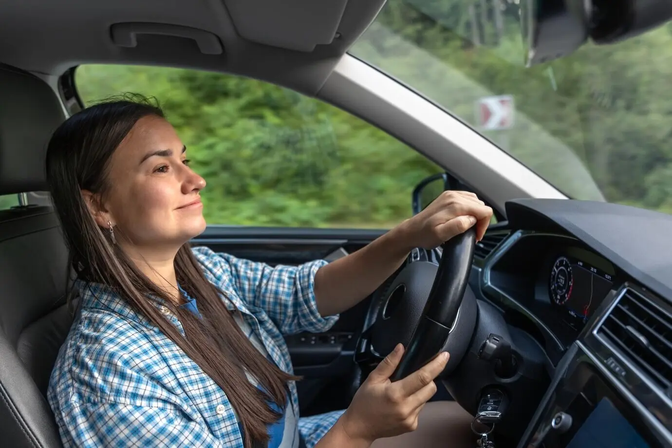 Сосредоточенная женщина ведет автомобиль по горной дороге, видны салон автомобиля и приборная панель.